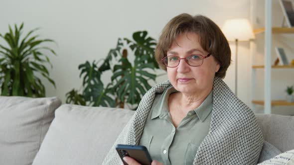Beautiful Old Lady Looking in Camera Sitting in the Living Room with Phone alt