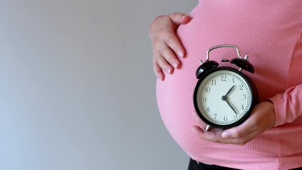 Pregnant young woman holds an alarm clock as a symbol of the imminent birth of her child.