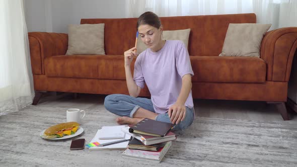 A Female Student Studies at Home with Textbooks Solves a Problem in a Notebook Sitting on the Floor