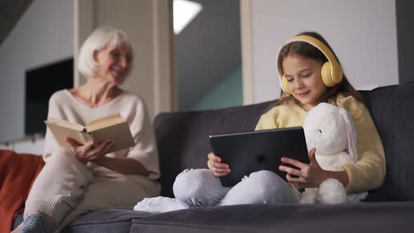 Girl looking at tablet and talking with grandmother reading book alt
