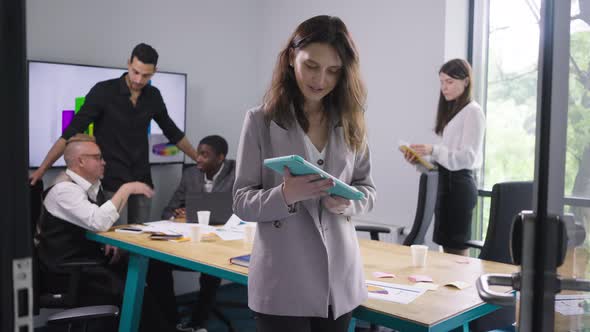 Smiling Satisfied Caucasian Woman Posing with Tablet in Office with Colleagues at Background alt