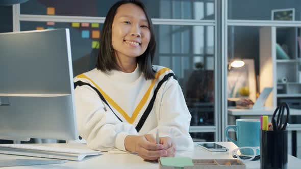 Portrait of Joyful Asian Employee Smiling Sitting at Computer Desk in Office alt