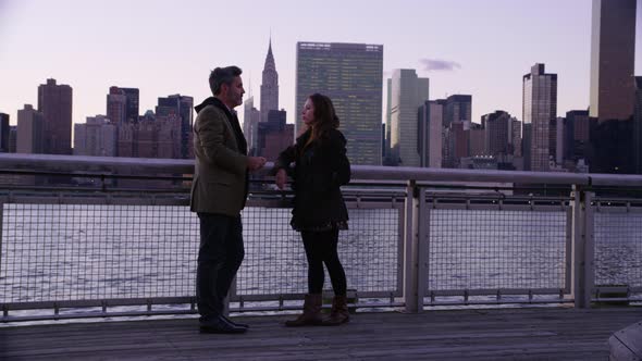 Couple in New York City stand by river talking with skyline in background