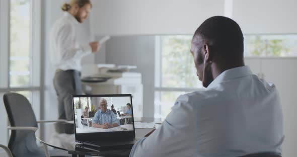 Back View of African Employee Sitting at Desk in Office Having Video Chat with Boss on Laptop alt