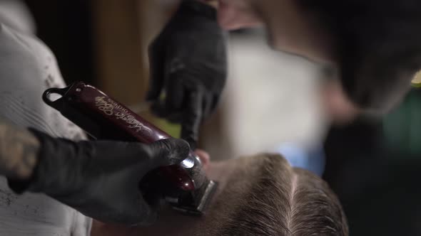 Tattoed man dressed in black T-shirt and cap, wearing glasses cutting client's hair with hairclipper alt