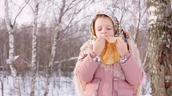 Cute girl in a traditional Russian headscarf with bagels eats pancake on winter background. alt