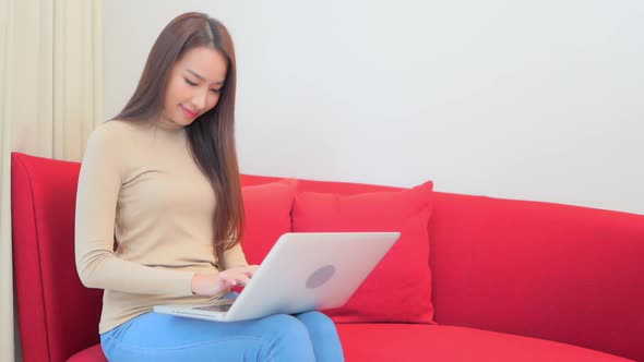 Asian woman in 30th typing on laptop computer holding it on laps sitting on red home sofa alt