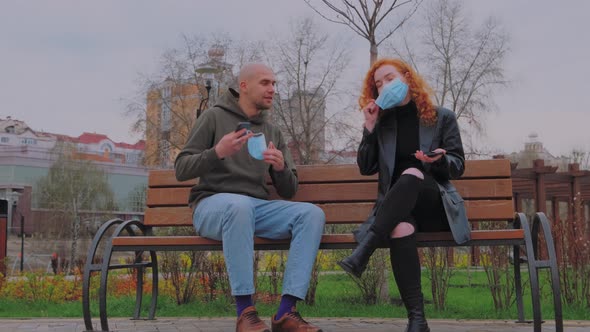 Man and Woman Sitting on Park Bench Take Off Their Protective Masks and Begin to Communicate alt