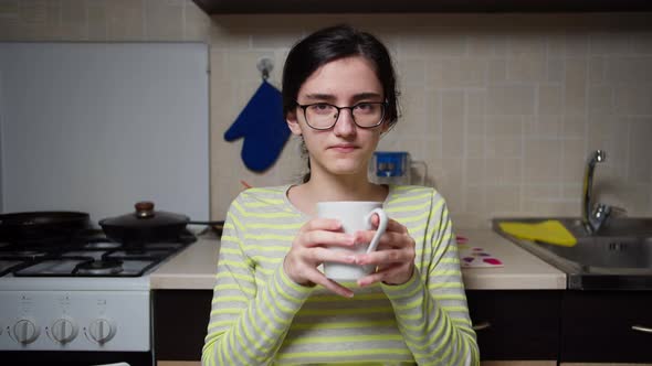 Girl with Glasses Is Sitting at the Kitchen Table Drinking a Hot Drink From a White Ceramic Mug alt