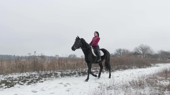 Young Brunette Woman Rides a Beautiful Black Horse on a Field or Snowcovered Farm in Winter alt