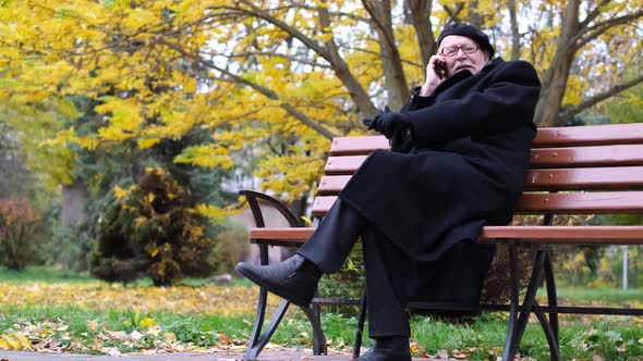 A French Senior is Talking on a Smartphone He is Sitting in a Park on a Bench alt
