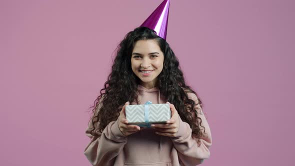 Portrait of Happy Young Woman Wearing Party Hat Shaking Gift Box on Pink Background alt