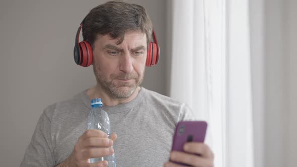 Positive Middle Aged Man in Headphones Drinking Refreshing Water and Listening to Playlist  alt
