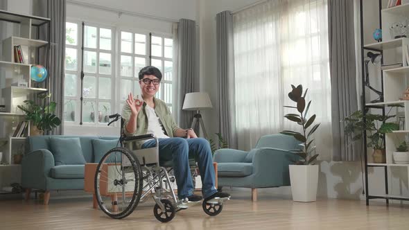 Asian Man Sitting In A Wheelchair Smiling And Showing Ok Sign In Living Room alt