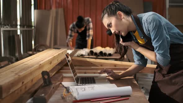 Young Asian female carpenter using laptop computer while talking to customer on the phone alt