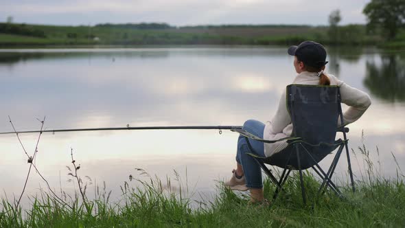 Woman with a Fishing Rod Sitting on a Chair By the Lake alt