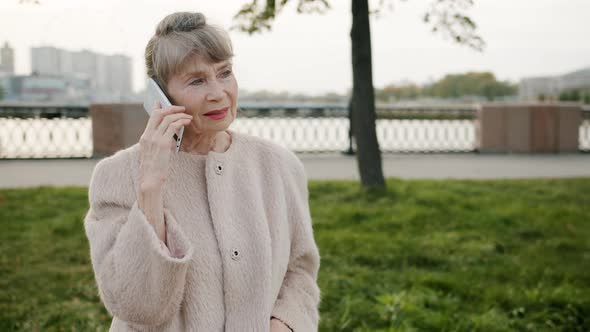 Portrait of Aged Greyhaired Woman Speaking on Mobile Phone Standing in City Street alt