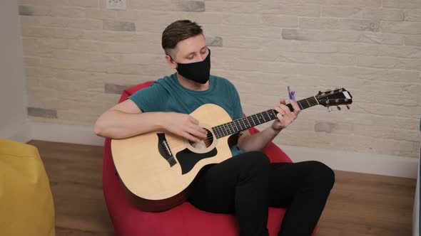 Young Man with a Black Medicinal Mask on Face Is Playing at the Guitar While Is Sitting Comfortably alt