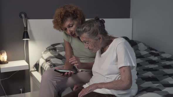 An Elderly Woman Looks at a Smartphone, with Her Adult Daughter at Bedroom. Daughter Teaching Senior alt