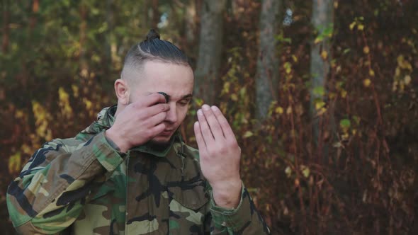 The Soldier in Camouflage Smears Soot on Face at a Forest alt