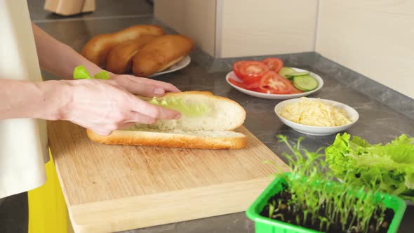 Hands spread lettuce leaves on a sandwich bun on a wooden cutting board.