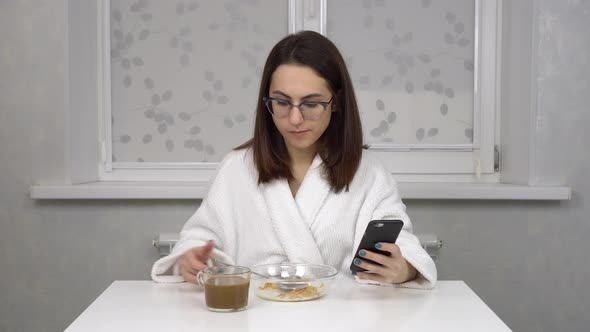 A Cheerful Young Woman in a White Coat Eats Cereals and Drinks Coffee and Uses a Smartphone alt