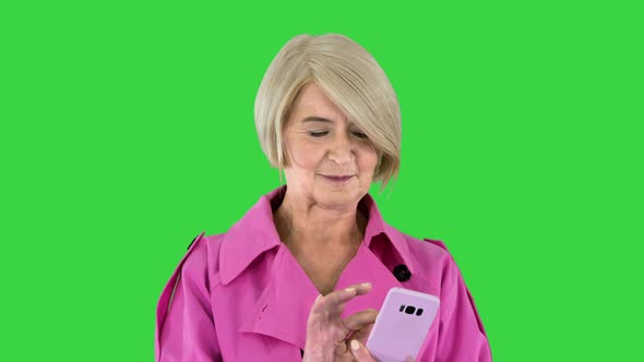 Cheerful Senior Lady in Pink Trench Coat Using Her Smartphone on a Green Screen Chroma Key alt