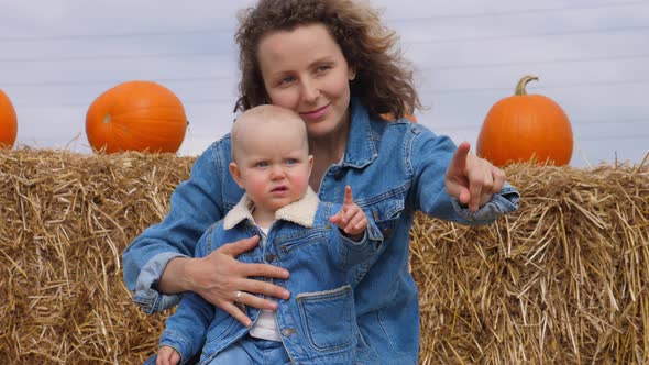 Portrait of Caucasian Mom and Baby in Matching Denim Looks Sitting on a Hay Bale Pointing at alt