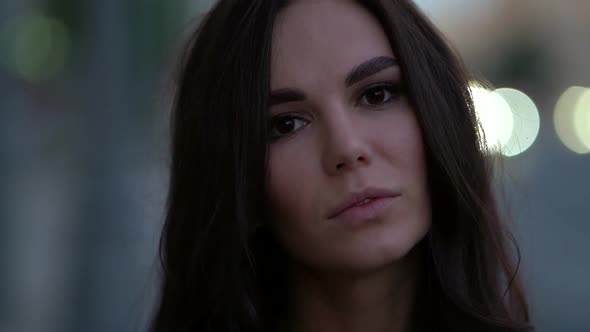 Close-up of a Young Brunette's Face Against a Background of Blurry Lights. She Looks at the Camera alt