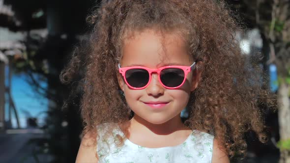 Close-up Portrait of a Beautiful Little Girl in Pink Glasses, Cute Smiling, Looking at the Camera alt