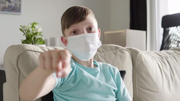 A Young Boy in a Face Mask Points at the Camera and Nods As He Sits on a Couch at Home alt