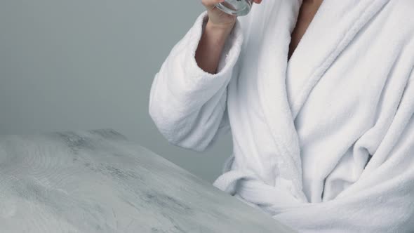 Thirsty smiling 35s woman in white bathrobe drinking water