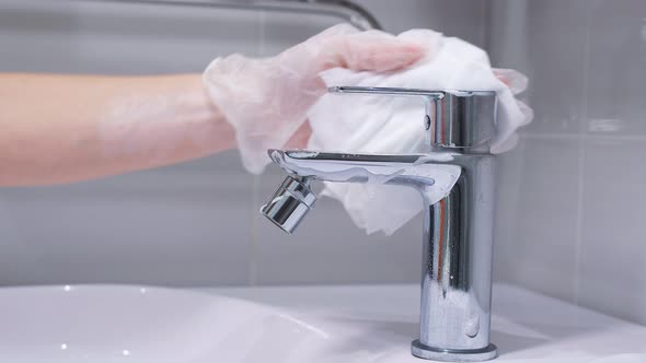 Woman's Gloved Hand Cleans a Stainless Steel Water Tap in the Bathroom with Detergent. Hygiene in alt