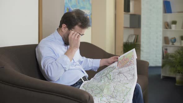 Male Traveler Sitting on Sofa and Looking at Map, Choosing Route for Vacation alt