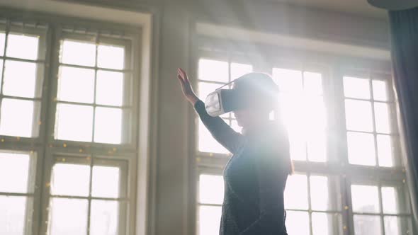 Young Woman Is Using Virtual Reality Glasses Indoors with Large Panoramic Window in Background alt