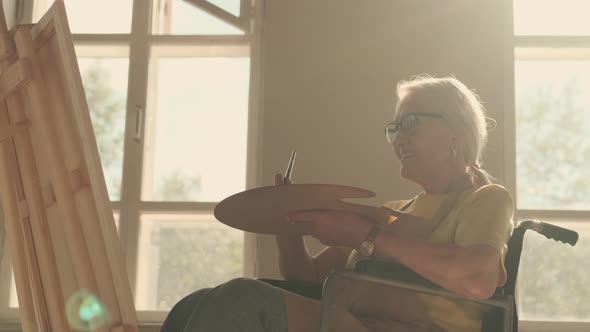 Senior Woman Disabled Artist Paints a Picture While Sitting in a Wheelchair alt