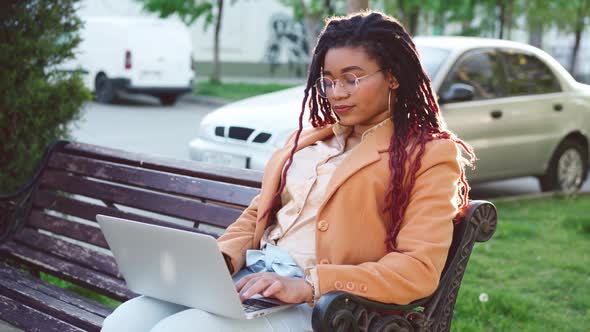 African American Woman Sitting on a Bench in City and Using Laptop