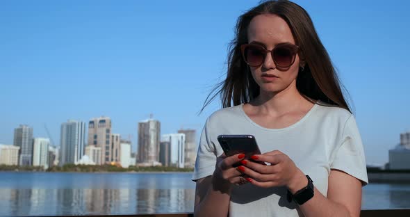Beautiful Girl with Long Hair in Sunglasses Using Smartphone App at Sunset River Quay Near alt