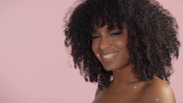 Mixed Race Black Woman with Curly Hair Covered By Crystal Makeup on Pink Background in Studio