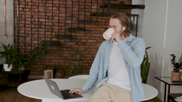 An Attractive Young Employee Having a Break Scrolling His Lapyop and Drinking Coffee alt