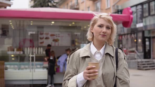Young Blonde Business Woman in a Beige Trench Coat and Vest White Blouse Stands with a Disposable alt