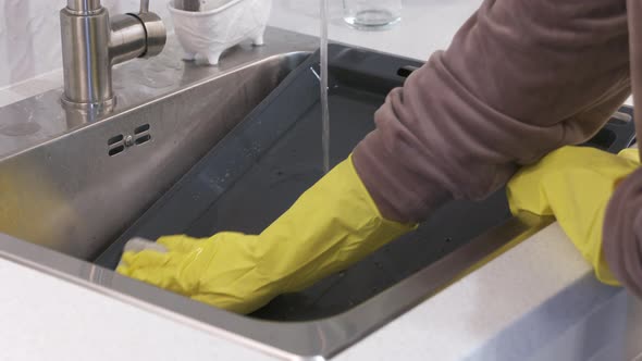 Woman Wearing Rubber Gloves Cleaning Oven Baking Sheet in Kitchen Sink Under Running Water alt