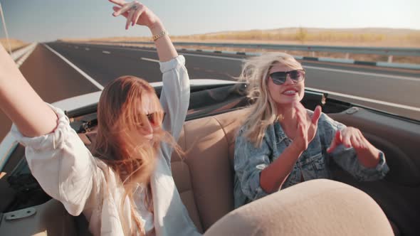 Happy cheerful female friends are driving in car with an open roof on highway, they are waving hands alt