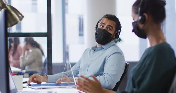 Diverse male and female colleague in face masks and headsets talking at desk with sneeze shield alt