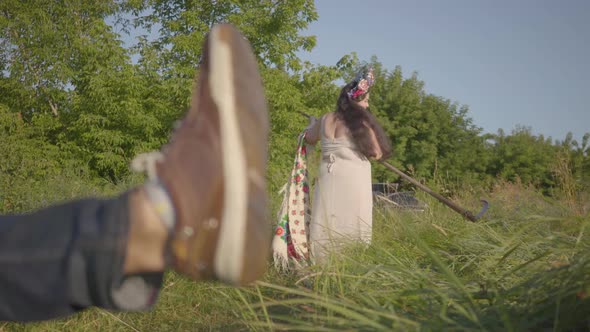 Foot of a Young Man Resting on the Ground in the Foreground. Plus Size Woman Finishing To Mow the alt
