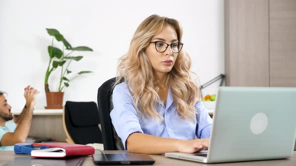Freelancer Woman Working on the Computer While Her Husband Is Watching a Ball Game in the Background alt