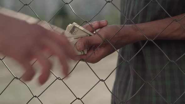 Close-up of Male African American and Caucasian Hands Passing Money and Pills Through Mesh Fence alt