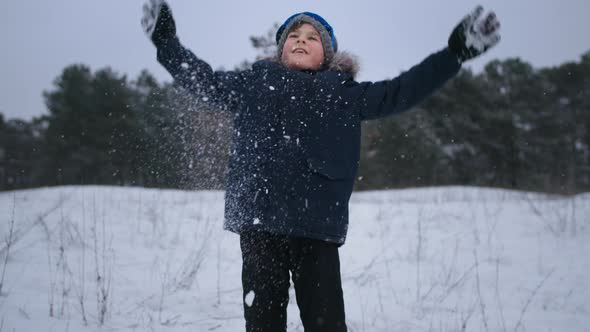Cheerful Winter Joyful Male Child in Warm Winter Clothes Throws Snow Background of Trees in Forest