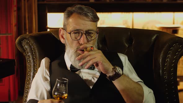 Noble Man Sits in Armchair with Cigar and Glass of Whiskey