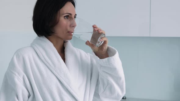 Thirsty smiling 35s woman in white bathrobe drinking water